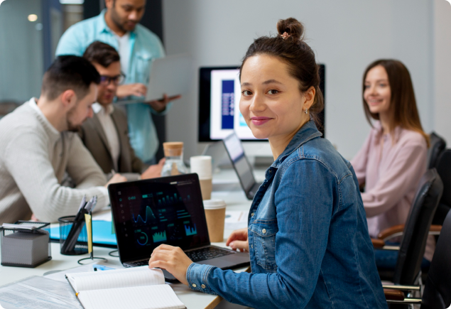 A woman in a denim jacket smiles at the camera while working on a laptop at a conference table with four colleagues, who are engaged in discussion and using laptops and documents.