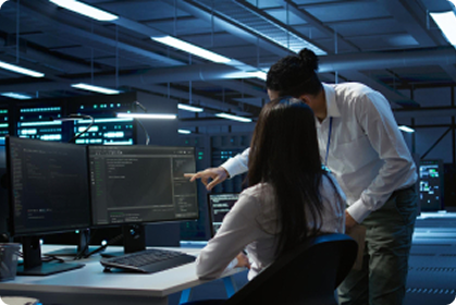 Two people in a modern, dimly lit office or data center are looking at computer monitors with code on the screens. One person is seated and typing, while the other stands and points at the monitor.