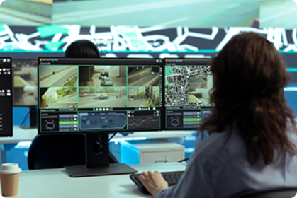 Two people sit at a desk with dual monitors displaying surveillance camera feeds and maps, monitoring traffic and city scenes. A takeaway coffee cup is on the desk. The background is modern and brightly lit.